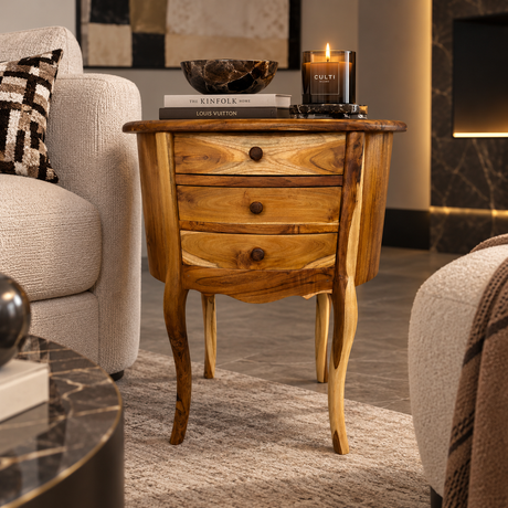 A rustic wooden three-drawer nightstand with curved cabriole-style legs, styled in a cozy living room next to a beige armchair with a lit candle on top.