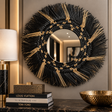 A round decorative wall mirror with a wide black and gold woven sunburst frame, displayed on a dark marble console against a beige wall with a lamp and books nearby.