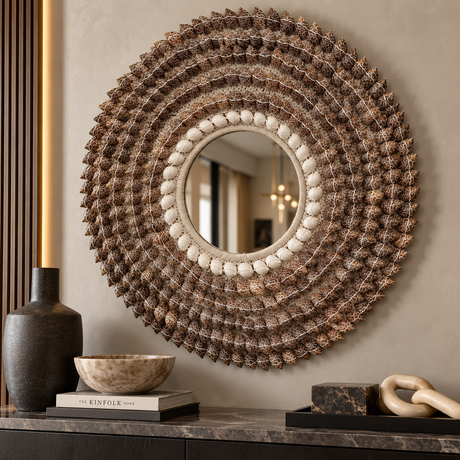 A large round decorative wall mirror with an intricate sunburst frame made of textured brown and beige elements, mounted on a beige wall above a console table with decorative objects.