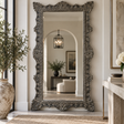 An ornate full-length floor mirror with a heavily carved, distressed silver-gray frame stands in a bright, modern entryway. The reflection shows a softly lit living room with an arched doorway, while the surrounding space features a large textured vase with greenery on the left and a marble console on the right.