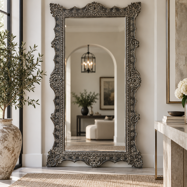 An ornate full-length floor mirror with a heavily carved, distressed silver-gray frame stands in a bright, modern entryway. The reflection shows a softly lit living room with an arched doorway, while the surrounding space features a large textured vase with greenery on the left and a marble console on the right.