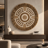 A large circular woven wall hanging with a geometric pattern and frayed fringe, displayed on a beige wall above a dark wood console in a modern living room setting.
