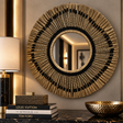 A large round wall mirror with a sunburst frame made of gold-toned rods and black accents, displayed in an elegant interior setting above a marble surface with books and a bowl.